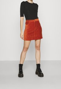 Mini-jupe en velours côtelé orange avec fermeture éclair avant, deux poches latérales et une coupe ajustée, associée à un haut noir à manches courtes et des bottes noires.