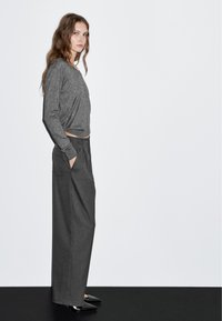 Grijze longsleeve top met een ontspannen pasvorm, gecombineerd met high-waisted, gestreepte wijde broek en zwarte puntige hakken, gepresenteerd tegen een effen achtergrond.