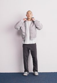 Doudoune gris clair avec col montant, fermeture éclair à l'avant et texture légèrement brillante. Associée à un t-shirt blanc et un pantalon foncé.