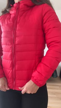 Άτομο που φοράει μια φωτεινή κόκκινη μπουφάν με φερμουάρ μπροστά, με τα χέρια να ρυθμίζουν το μπουφάν κοντά στη μέση, όρθιο σε εσωτερικό χώρο πάνω σε ξύλινο πάτωμα.