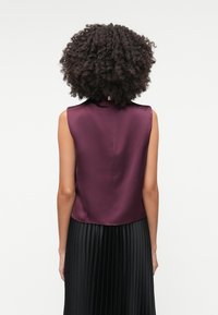 Top viola in satin senza maniche con colletto alto, caratterizzato da una texture liscia e una leggera curvatura nell'orlo, abbinato a una gonna nera plissettata.