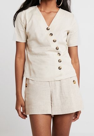 Vrouw draagt een beige korte mouwen V-hals top met diagonale knopen, gecombineerd met beige shorts met hoge taille en zijknopen.