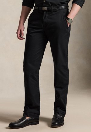 Homme portant un pantalon noir, une ceinture en cuir noire, une chemise noire aux manches retroussées, une montre noire et des mocassins noirs, debout avec une main dans la poche.