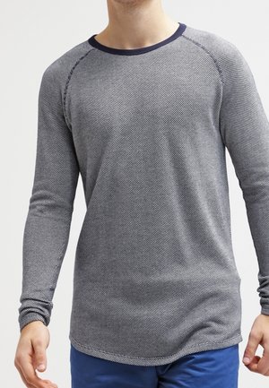 Man die een langemouwige, getextureerde grijze trui met marineblauwe rand en een blauwe broek draagt, staand tegen een effen achtergrond.