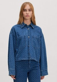 Blaue, taillierte Jeansjacke mit Knopfverschluss, spitzem Kragen und zwei Fronttaschen, die ein dezentes Texturmuster aufweist.