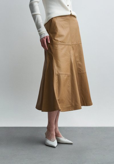 Femme portant une jupe midi en cuir couleur beige coupe trapèze, un top à manches longues blanc côtelé et des talons blancs à bout pointu, debout sur un sol gris.