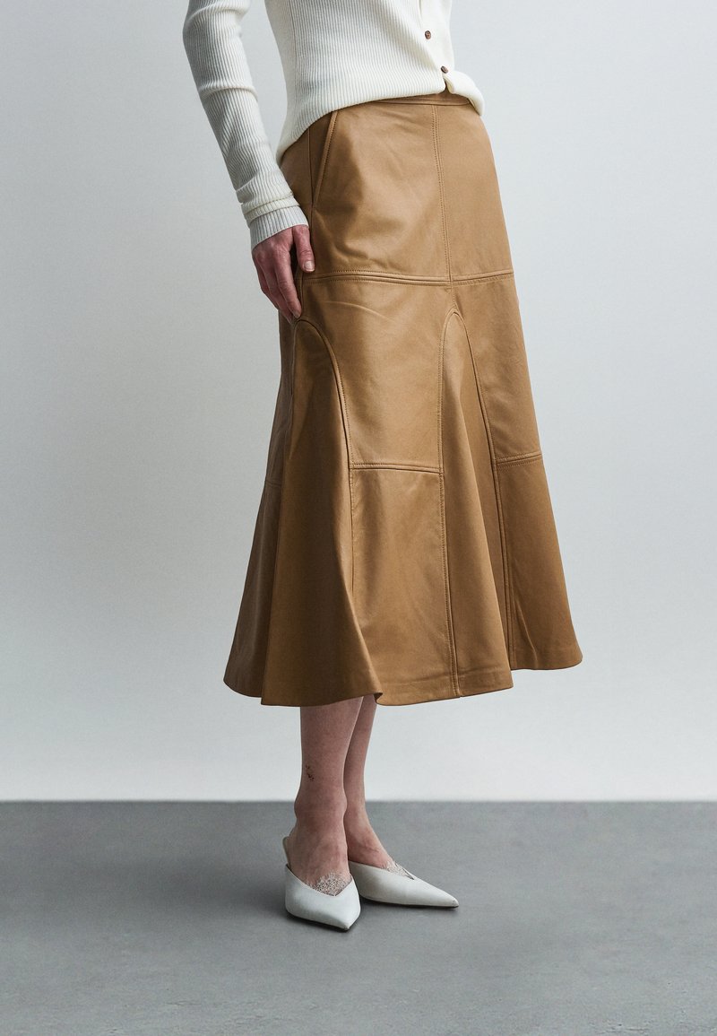 Γυναίκα που φοράει μπεζ δερμάτινη μίντι φούστα σε γραμμή Α, λευκό ριμπ μακρυμάνικο τοπ και λευκά μυτερά ψηλοτάκουνα, στέκεται σε γκρι πάτωμα.