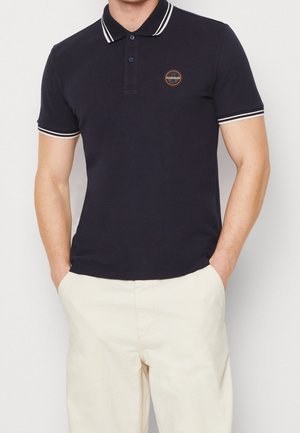 Mann trägt ein schwarzes Kurzarm-Poloshirt mit weißem Besatz am Kragen und an den Ärmeln, kombiniert mit hellbeigen Hosen, Hände in den Taschen.