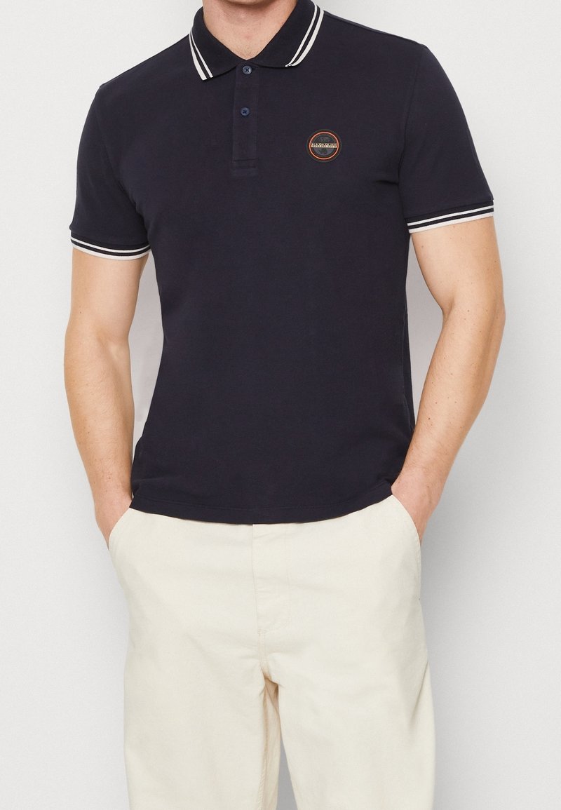 Homme portant un polo noir à manches courtes avec un bord blanc au col et aux manches, assorti à un pantalon beige clair, les mains dans les poches.