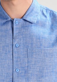 Närbild av en ljusblå skjorta med krage och bröstficka, bärs av en person som visar hals och övre bröst.