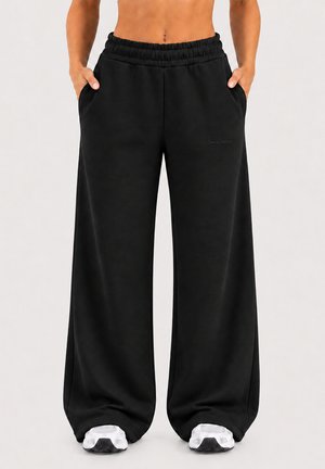 Persona con pantalones anchos negros y sueltos con cintura elástica y zapatillas blancas, de pie con las manos en los bolsillos, con un fondo claro.
