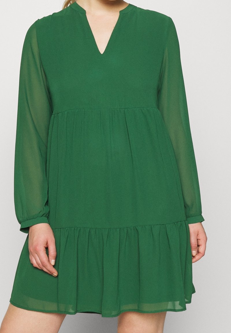 Zielona sukienka do kolan wykonana z lekkiego materiału, z długimi rękawami, dekoltem w serek, warstwowymi falbanami i luźnym fasonem.