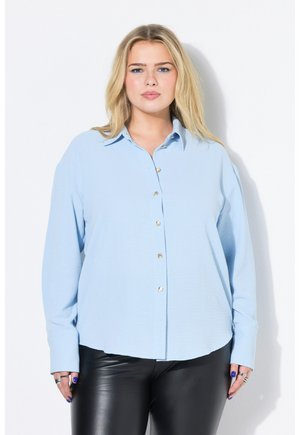 Blondynka w jasnoniebieskiej koszuli na guziki i czarnych skórzanych spodniach, stojąca na białym tle.