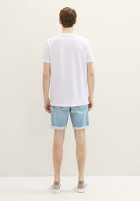 Camiseta de algodón blanca, pantalones cortos de mezclilla claros con puños doblados y zapatos grises sin cordones. Modelo visto desde atrás contra un fondo liso.