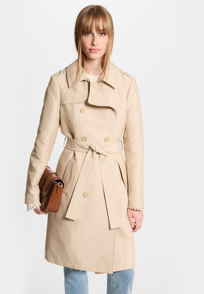 Beige trenchcoat met een dubbele knoopsluiting, strik taille, zijzakken en een brede kraag. Model houdt een bruine handtas vast.