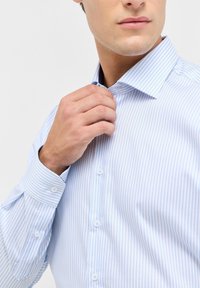 Uomo che regola il colletto di una camicia dress shirt a righe azzurre e bianche su uno sfondo semplice.