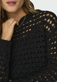 Schwarzer Strickpullover mit einem offenen Strickdesign, gerippten Bündchen und rundem Ausschnitt. Der Stoff hat ein strukturiertes, atmungsaktives Aussehen.
