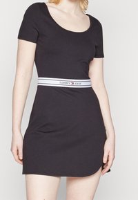 Musta lyhythihainen mekko, joka on valmistettu joustavasta kankaasta. Mekossa on pyöreä pääntie ja valkoinen joustava vyötärö, jossa on "TOMMY JEANS" logo.