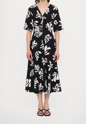 Mujer con vestido midi negro con patrones abstractos de hojas blancas, mangas cortas, detalle de lazo delantero y sandalias negras con tiras.