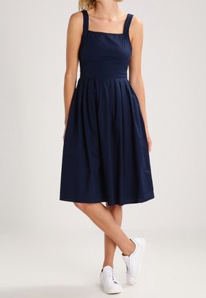 Frau trägt ein ärmelloses, knielanges, marineblaues Kleid mit quadratischem Ausschnitt und weißen Turnschuhen, steht mit gekreuzten Beinen.