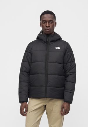 Homme portant une doudoune noire avec capuche et fermeture éclair, debout devant un fond clair uni.