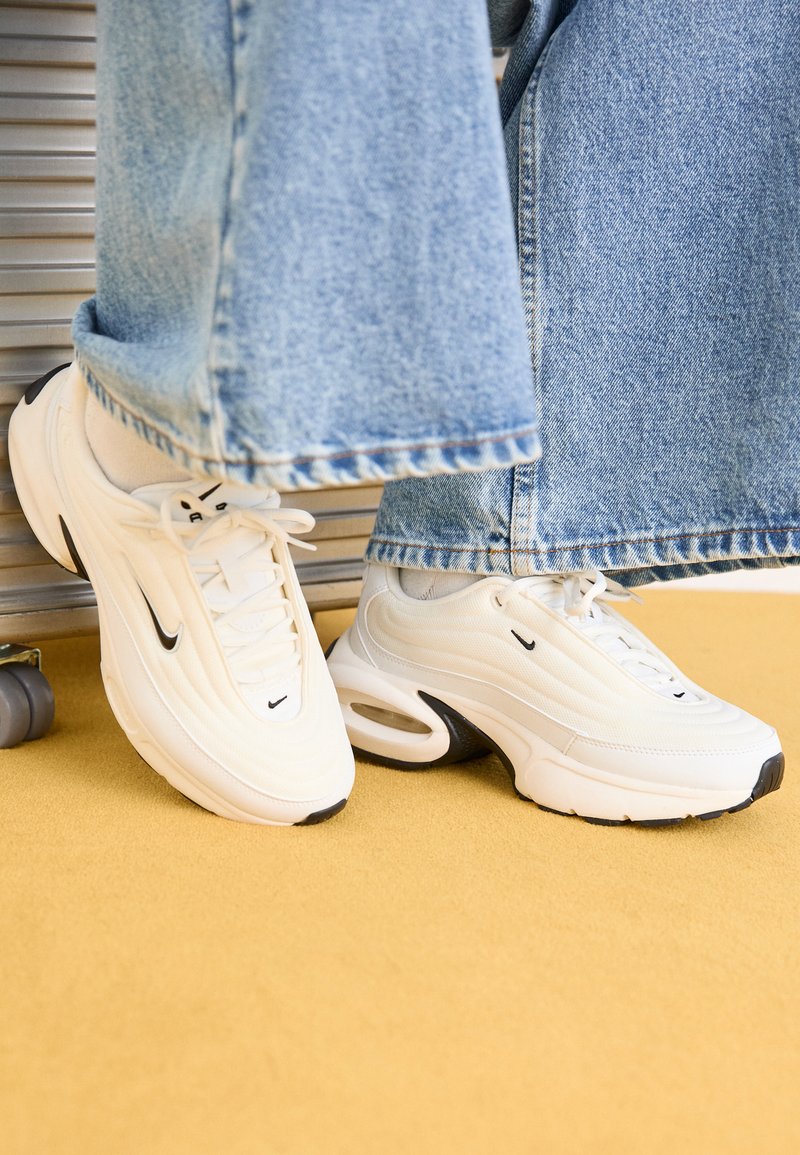Baltos Nike sportbačiai su tekstūruotu dizainu, juodais akcentais ir oro pagalvėlėmis. Pateikti po šviesiai mėlynomis džinsinėmis kelnėmis su nutrintais ilgiais.