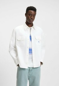 Hvid skjorte med knapper, to brystlommer, lange ærmer og en afslappet pasform iført over en blå og hvid grafisk T-shirt. Lyse denimbukser.