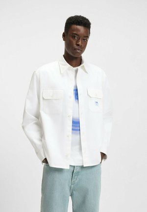 White button-up shirt with two chest pockets, long sleeves, and a relaxed fit worn over a blue and white graphic T-shirt. Light denim pants.