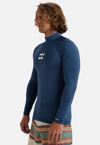 Herrarnas långärmade rash guard i marinblått med hög krage, som har en vit logo på bröstet och ett mjukt, elastiskt tyg.