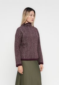 Woman wearing a maroon and white chevron-patterned sweater and olive green skirt, standing against a plain white background.