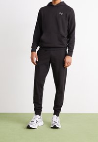 Sudadera negra con capucha y bolsillo delantero, combinada con pantalones deportivos a juego. Presenta un pequeño logotipo blanco en el pecho. Las zapatillas deportivas son blancas con detalles en negro.