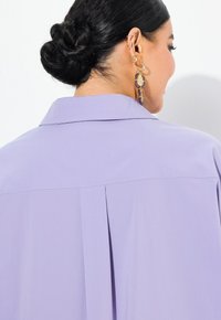 Světlolila blůza s límečkem, umocněná zadním skladem a jemnou texturou, doplněná zlatými a purpurovými náušnicemi.