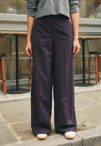 Brede, geruite broek in donkerblauw en bordeaux, gemaakt van zachte stof. Heeft een hoge taille en subtiele plooien aan de voorkant, gecombineerd met witte schoenen.