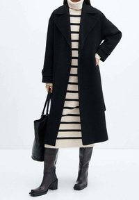 Manteau en laine noire avec de larges revers, porté par-dessus une robe-pull à rayures crème. Des bottes à talons marron montant jusqu'aux genoux et un sac fourre-tout noir complètent le look.