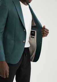 Blazer turchese con una vestibilità slim, realizzato in tessuto texturizzato. Presenta due bottoni e una fodera a righe chiare in contrasto.