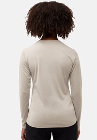 Persoon met krullend haar, gekleed in een beige overhemd met lange mouwen en een zwarte broek, weergegeven van achteren tegen een witte achtergrond.