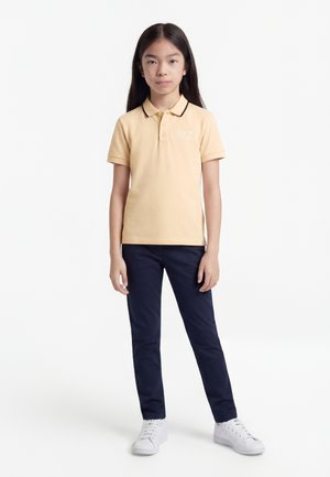 Meisje met lang donker haar, gekleed in een beige poloshirt, marineblauwe broek en witte sneakers, staand tegen een effen witte achtergrond.