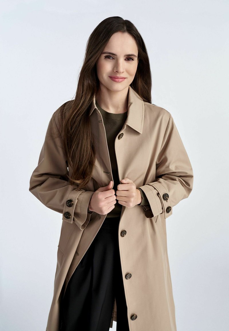 Beige Trenchcoat mit Knopfleiste, großen Knöpfen und Seitentaschen, kombiniert mit einem dunklen Pullover. Glatte Textur und taillierte Passform.