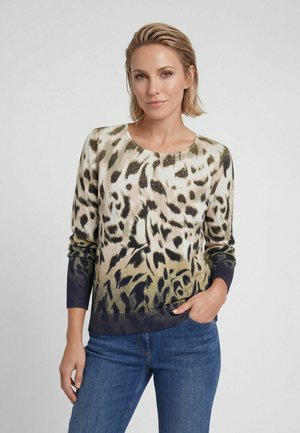 Frau mit kurzen blonden Haaren, die einen langärmeligen Pullover mit Tiermuster und blaue Jeans trägt, steht mit einer Hand in der Tasche vor einem schlichten Hintergrund.