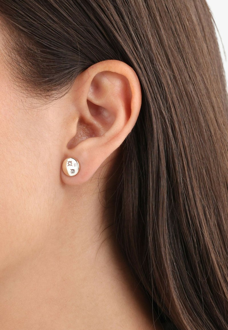 Orecchini a perno in oro con una superficie liscia e rotonda, caratterizzati da una piccola stella e un design circolare. Esibiti contro l'orecchio di un modello.