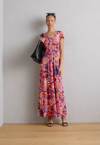 Robe maxi florale avec des motifs rose, orange et violet ; taille cintrée, manches courtes. Sac fourre-tout noir et sandales à lanières.