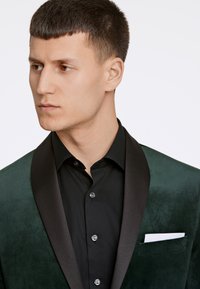 Blazer verde in velluto con colletto a scialle in satin nero, indossato sopra una camicia nera con bottoni. Un fazzoletto bianco è visibile nella tasca.
