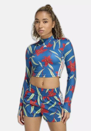 Donna che indossa un top corto blu a maniche lunghe e pantaloncini abbinati con stampa floreale rossa e strisce bianche, in piedi davanti a uno sfondo semplice.