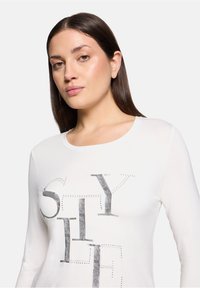 Langärmliges weißes T-Shirt mit silbernem Metallic-Druck, der "STYLE" mit Nietenakzenten buchstabiert; glattes Gewebe mit taillierter Passform.