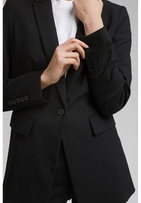 Czarna marynarka wykonana z gładkiego materiału, z zapięciem na jeden guzik, dopasowanym kroju i mankietami z detalami w postaci czterech guzików. Kieszenie po bokach.