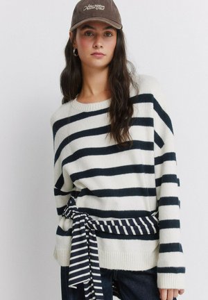 Vrouw met lang donker haar die een bruine pet, een witte trui met zwarte horizontale strepen en een gestreepte stoffen riem die in de taille is gestrikt, draagt.