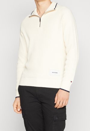 Man wearing a cream-colored ribbed sweater with a half-zip collar and black pants with side pockets, standing against a plain background.