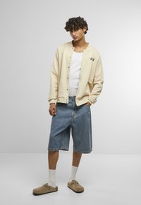 Béžová mikina na gombíky, biele tričko bez rukávov pod ňou, svetlomodré denimové kraťasy, béžové dreváky, biele ponožky do polovice lýtok, textúrovaná látka.