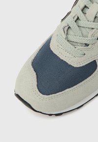 Jasnoszary zamszowy i ciemnoniebieski siateczkowy sneaker z fakturowaną powierzchnią, sznurowadłem i gumową podeszwą. Charakteryzuje się przeszyciami oraz zaokrąglonym kształtem noska.