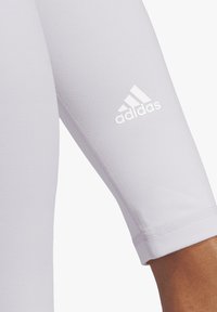 Jasnofioletowa koszulka sportowa z rękawami o długości trzech czwartych, z białym logo Adidas na rękawie i gładką, elastyczną fakturą materiału.